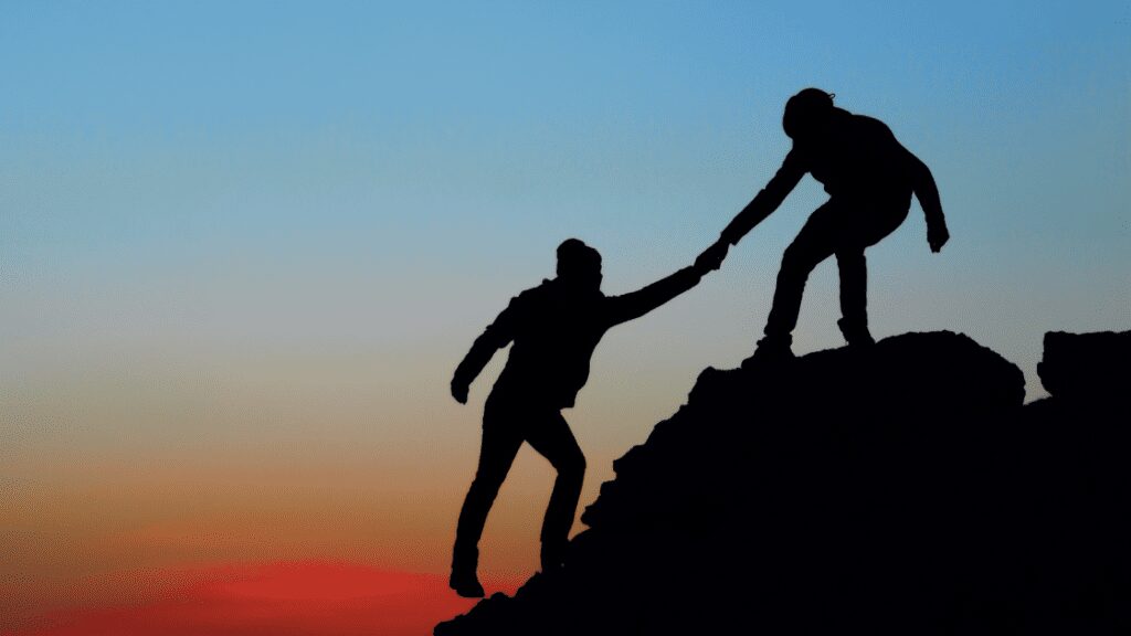 man helping another man up a mountain at sunset